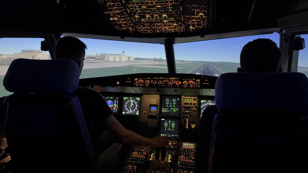 Zwei Personen auf den Pilotensitzen des Airbus-Cockpits im Simulator, vor ihnen die Startbahn, darüber das beleuchtete Overhead-Panel